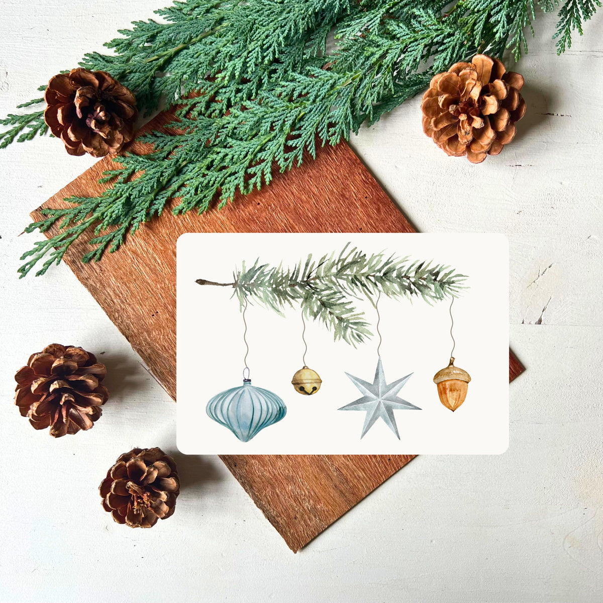 Tannenzweig an dem verschiedene Dekoelemente hängen (Weihnachtskugel in türkis, Glöckchen, Weihnachtsstern und Eichel) auf einer Postkarte von foodigence fotografiert vor Tannengrün