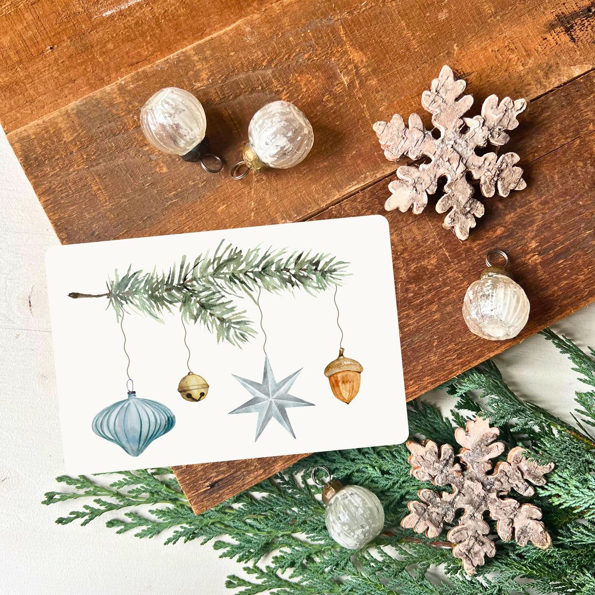 von foodigence designte Weihnachtskarte mit türkisen Weihnachtselementen, die an einem Tannenzweig hängen aquarell-gemalt abgebildet ist die Postkarte vor einem Holzbrett mit Weihnachtsdeko