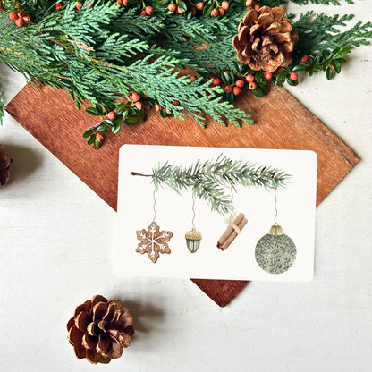 Weihnachtskarte von foodigence mit Tannenzweig an dem verschiedene Dekoelemente hängen (Lebkuchen, Porzellan-Eichel, Zimtstangen und grüne Weihnachtskugel) dekoriert mit Tannengrün
