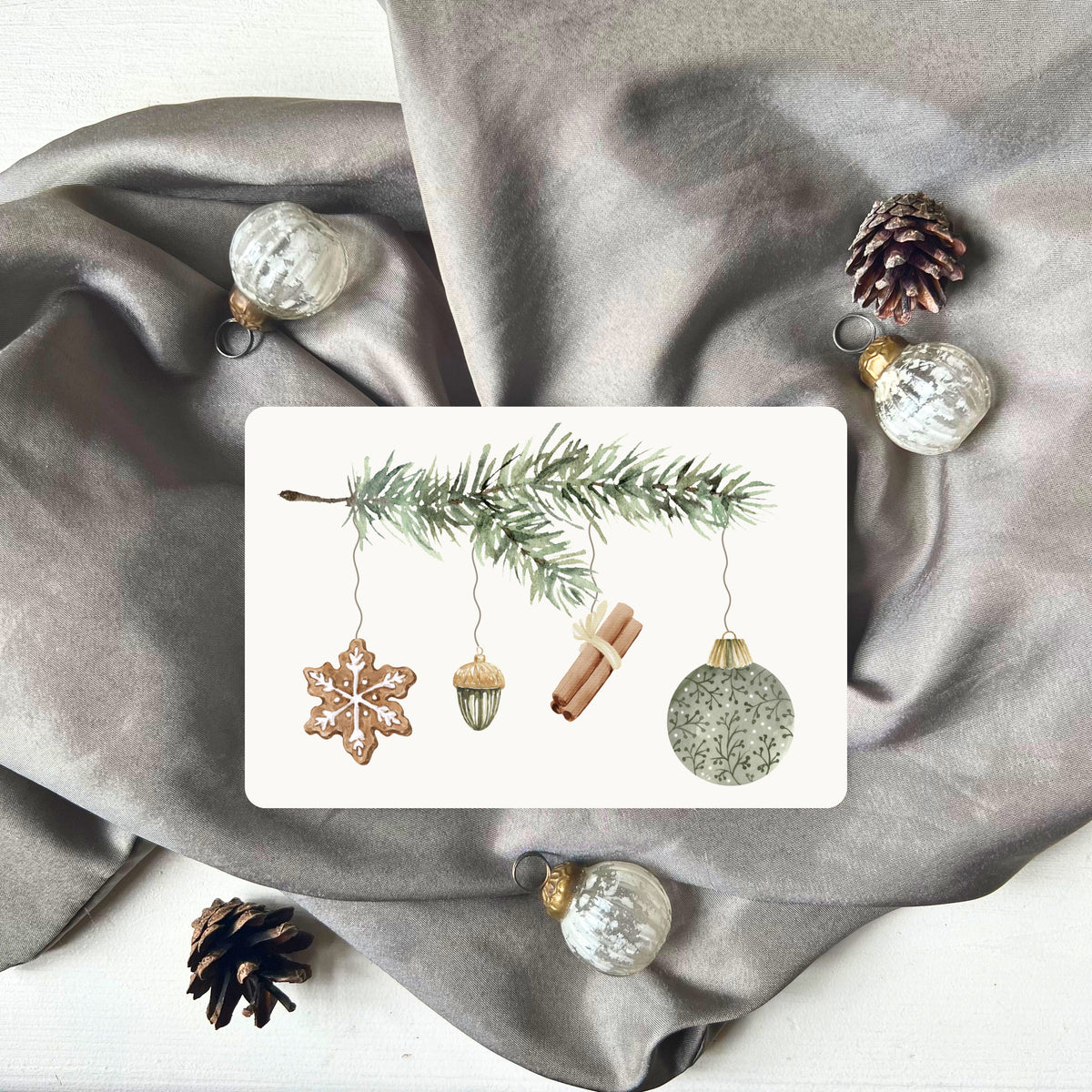 Weihnachtspostkarte von foodigence mit grünen Weihnachtselementen in aquarell-Design vor einem grauen Samttuch abgebildet 