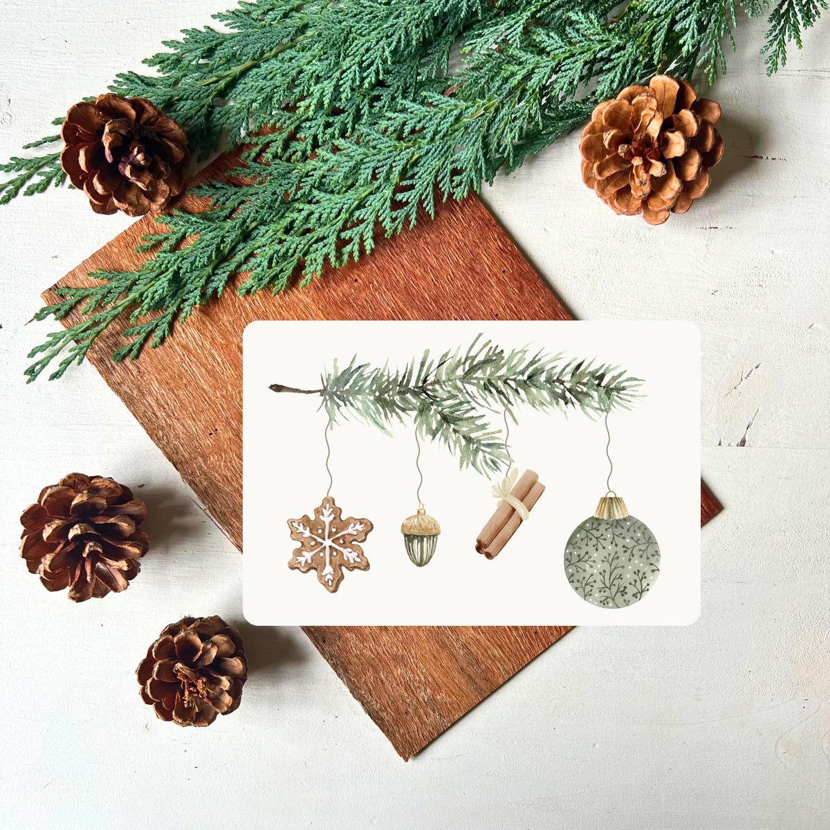 Tannenzweig an dem verschiedene Dekoelemente hängen (Lebkuchen, Porzellan-Eichel, Zimtstangen und grüne Weihnachtskugel) auf einer Postkarte von foodigence fotografiert vor Tannengrün