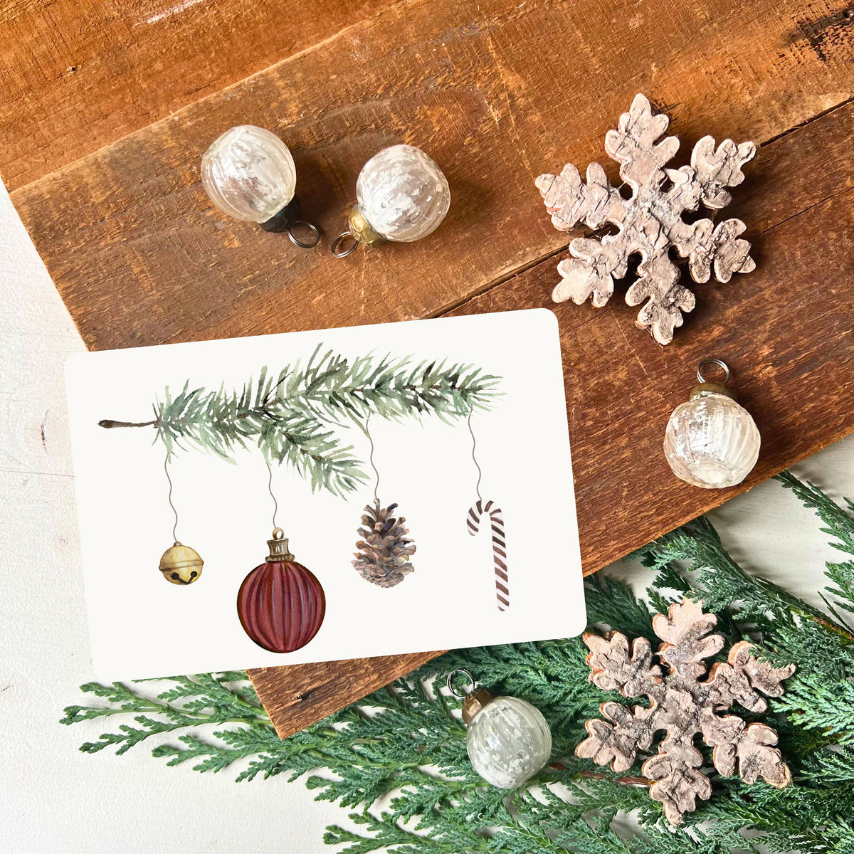von foodigence designte Weihnachtskarte mit dunkelroten Weihnachtselementen, die an einem Tannenzweig hängen aquarell-gemalt abgebildet ist die Postkarte vor einem Holzbrett mit Weihnachtsdeko