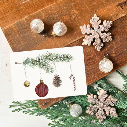 von foodigence designte Weihnachtskarte mit dunkelroten Weihnachtselementen, die an einem Tannenzweig hängen aquarell-gemalt abgebildet ist die Postkarte vor einem Holzbrett mit Weihnachtsdeko