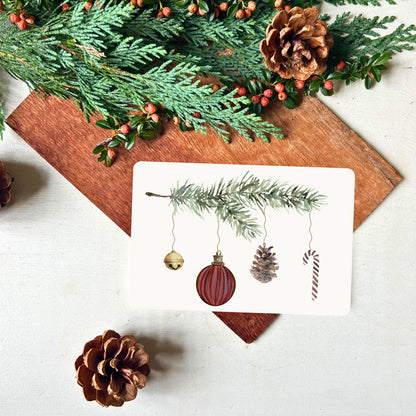 Weihnachtskarte von foodigence mit Tannenzweig an dem verschiedene Dekoelemente hängen (Glöckchen in gold, Weihnachtskugel in dunkelrot, Tannenzapfen und Zuckerstange) dekoriert mit Tannengrün