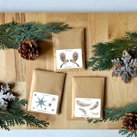 Übersicht Vogelfutter für heimische Vogelarten eingepackt mit winterlichem Etikett von foodigence - 3 Designs in der Übersicht (Abbildung mit Vögeln, Abbildung mit Sternen, Abbildung mit Federn) abgebildet sind die Sets vor winterlciher Deko